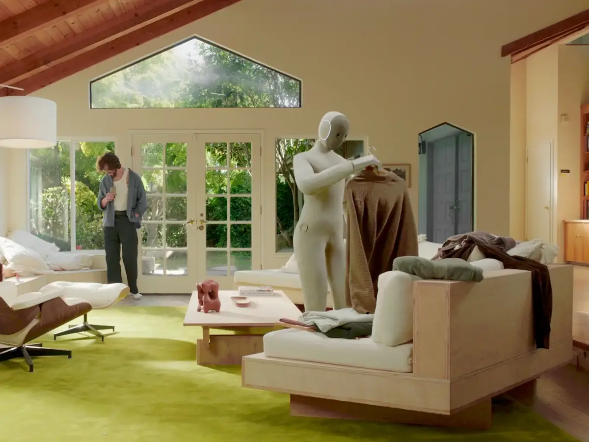 1X NEO humanoid robot hanging up a sweater in a sunny living room while a man stands by the garden doors (1X company photo)